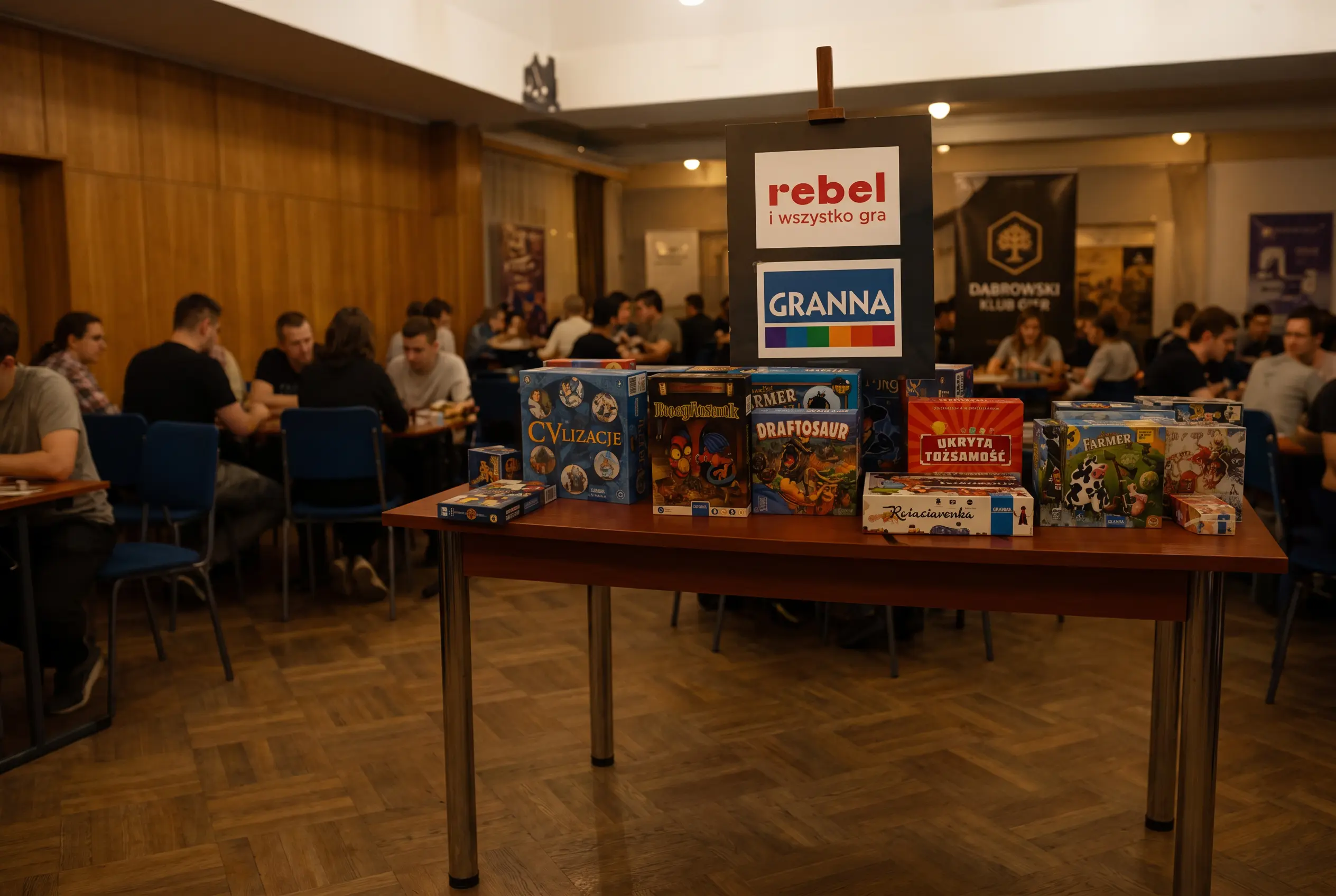 Stoisko sponsorów podczas wydarzenia Dąbrowskiego Klubu Gier Planszowych z grami Rebel i Granna oraz uczestnikami grającymi w planszówki.